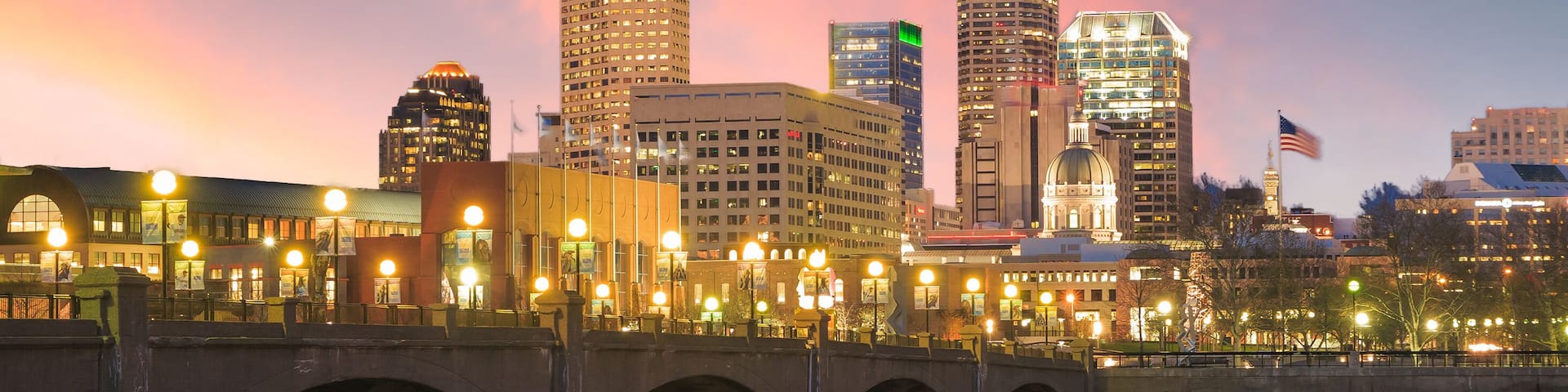A city skyline at dusk with illuminated buildings, a bridge, and a river reflecting lights.