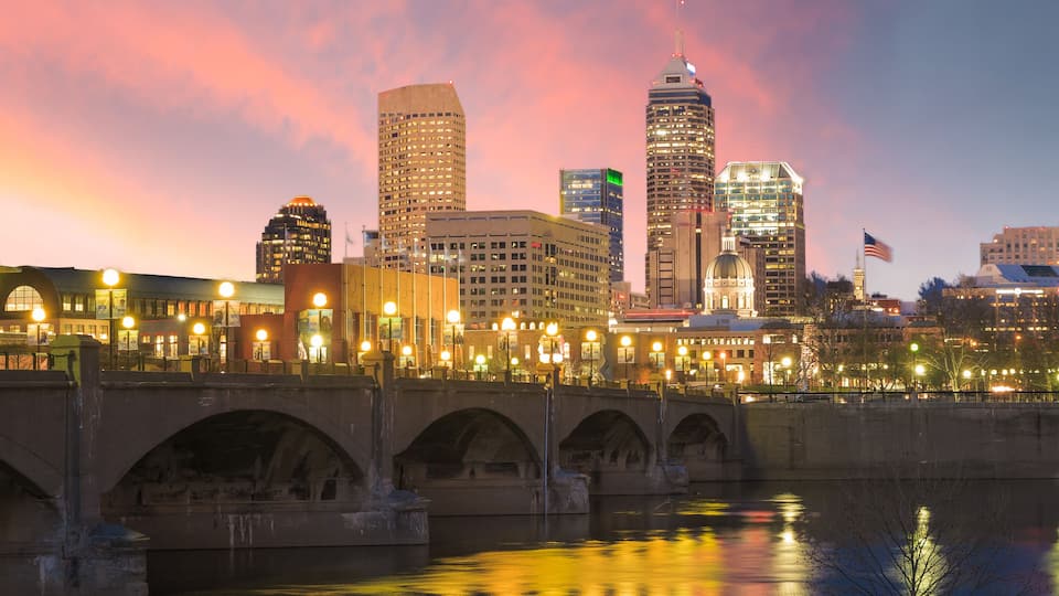 A city skyline at dusk with illuminated buildings, a bridge, and a river reflecting lights.
