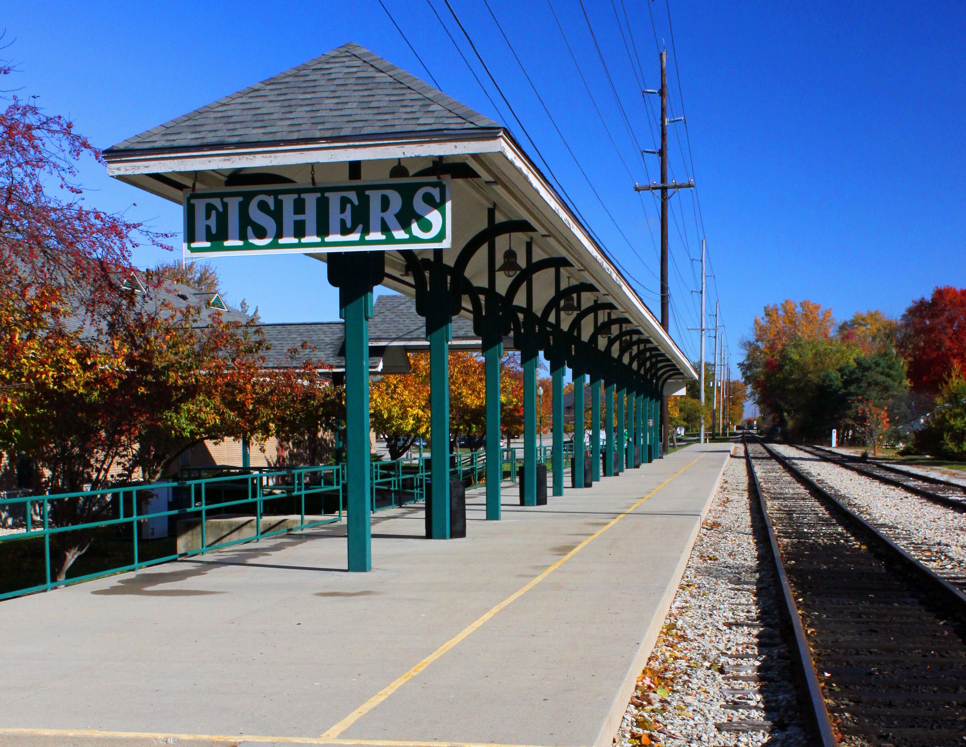 A train station platform with a 'FISHERS' sign, green pillars, and autumn-colored trees.
