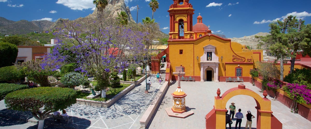 A church with a bell tower, surrounded by a plaza with people, trees, and mountains in the background.