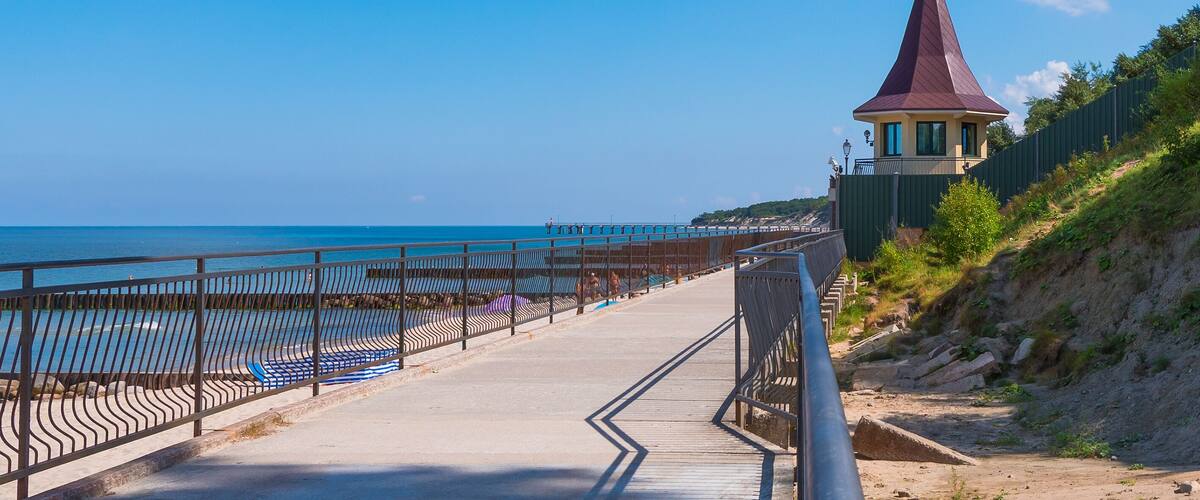 A coastal walkway with a red-roofed tower, a clear blue sky, and the ocean.