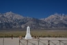 A white monument with inscriptions in a foreign script, surrounded by a rustic fence, set against a backdrop of rugged mountains and a clear blue sky.