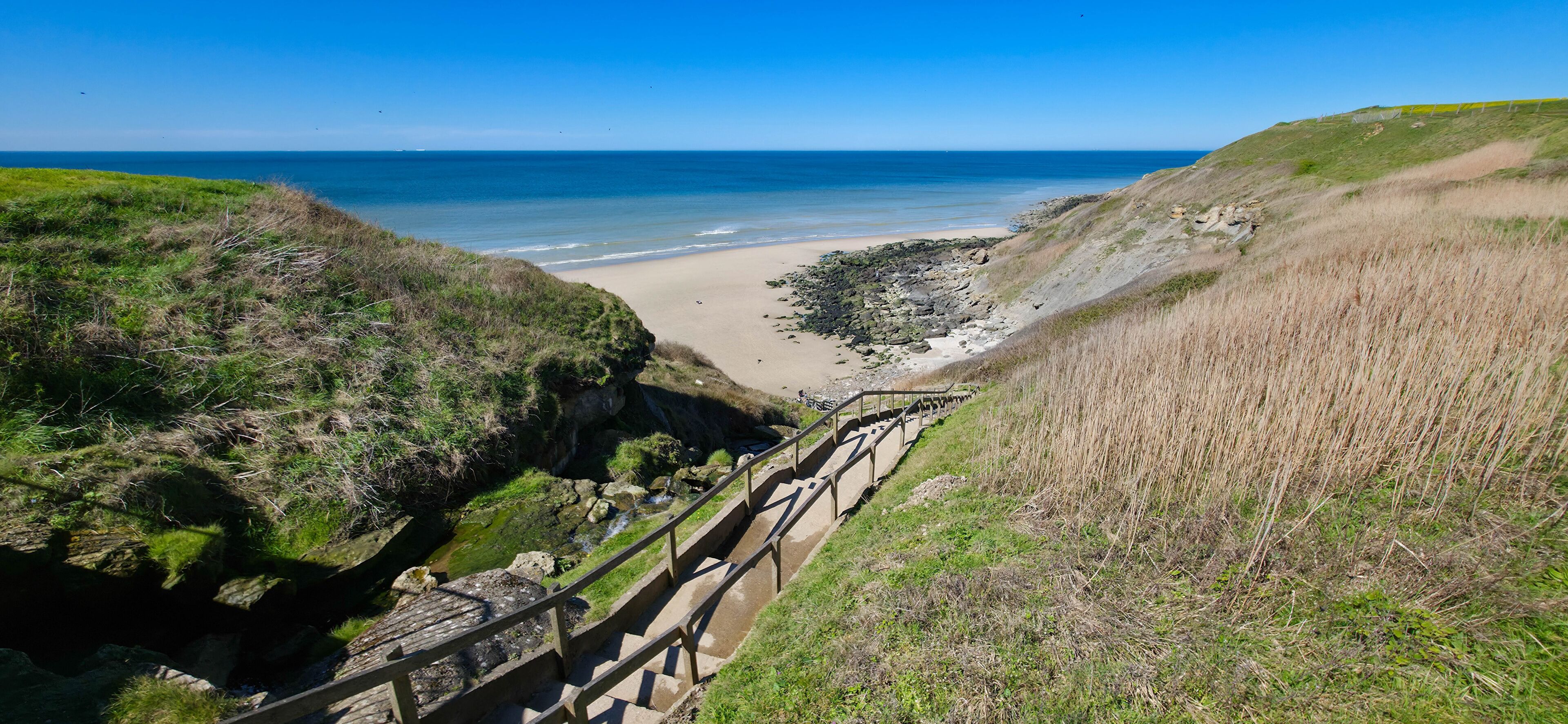 A coastal landscape with a sandy beach, grassy cliffs, and a wooden staircase leading down to the shore.