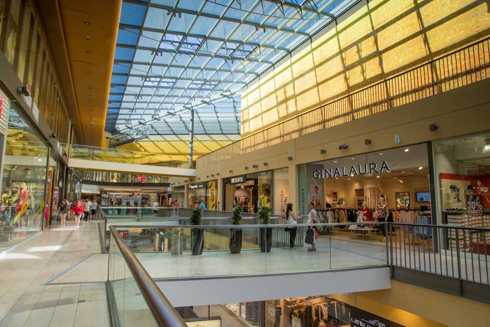 Un centre comercial modern amb sostre de vidre, diverses plantes i nombroses botigues.