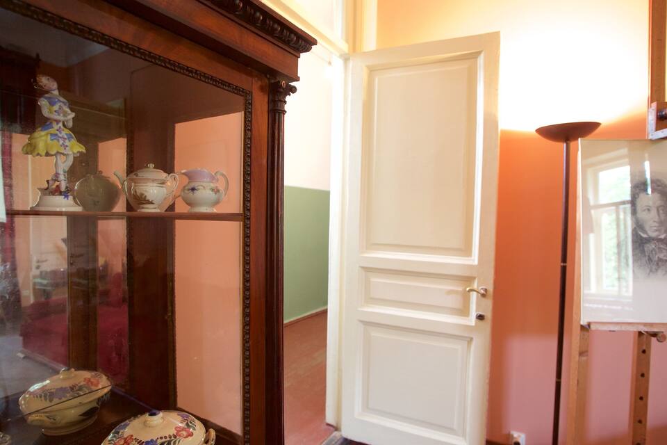 Una stanza con un armadietto in legno che espone servizi da tè in porcellana, una porta bianca e un ritratto incorniciato appeso al muro.