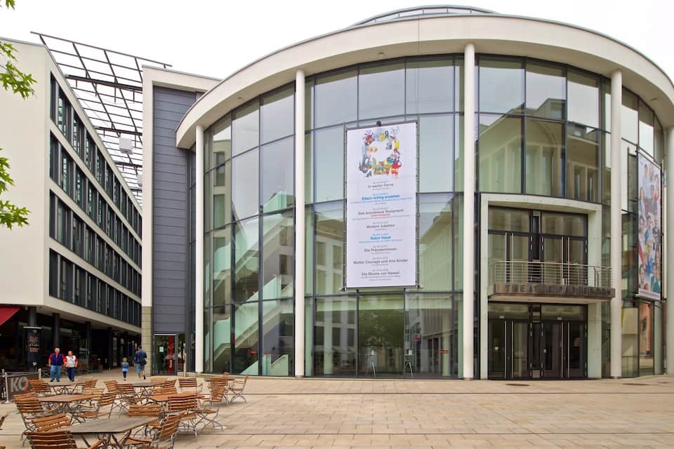Een modern gebouw met grote glasramen en een gebogen gevel. Er zijn buitenzitplaatsen met tafels en stoelen voor het gebouw.