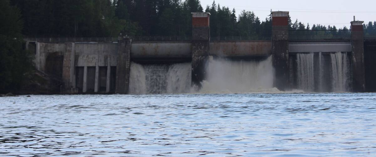 A dam with water flowing over it, a bridge, and a forested area in the background.