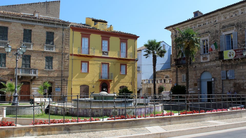 A yellow building with balconies, a fountain, and red flowers in a square.