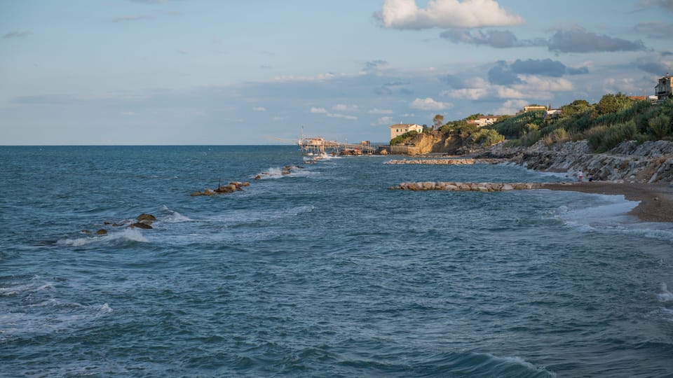 A coastal scene with waves crashing onto a rocky shore, a lighthouse, and buildings on a hillside.