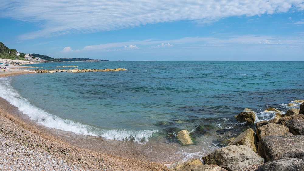 A pebble beach with clear water, a rocky breakwater, and a distant shoreline with buildings.