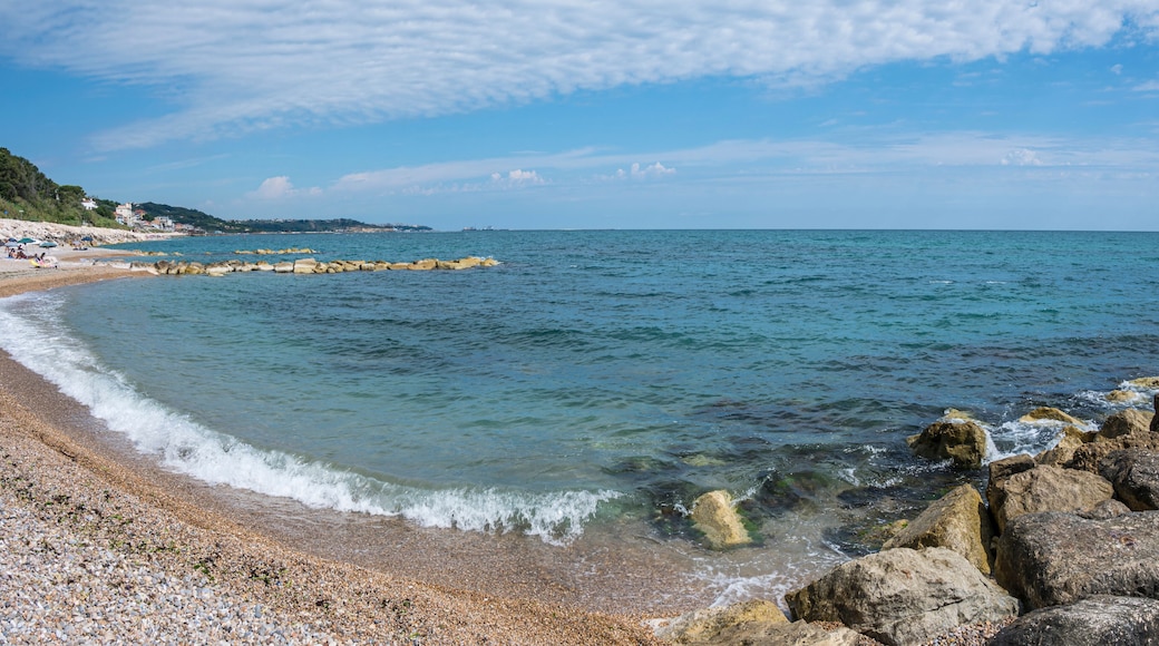 A pebble beach with clear water, a rocky breakwater, and a distant shoreline with buildings.