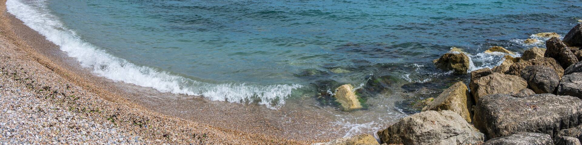 A pebble beach with clear water, a rocky breakwater, and a distant shoreline with buildings.