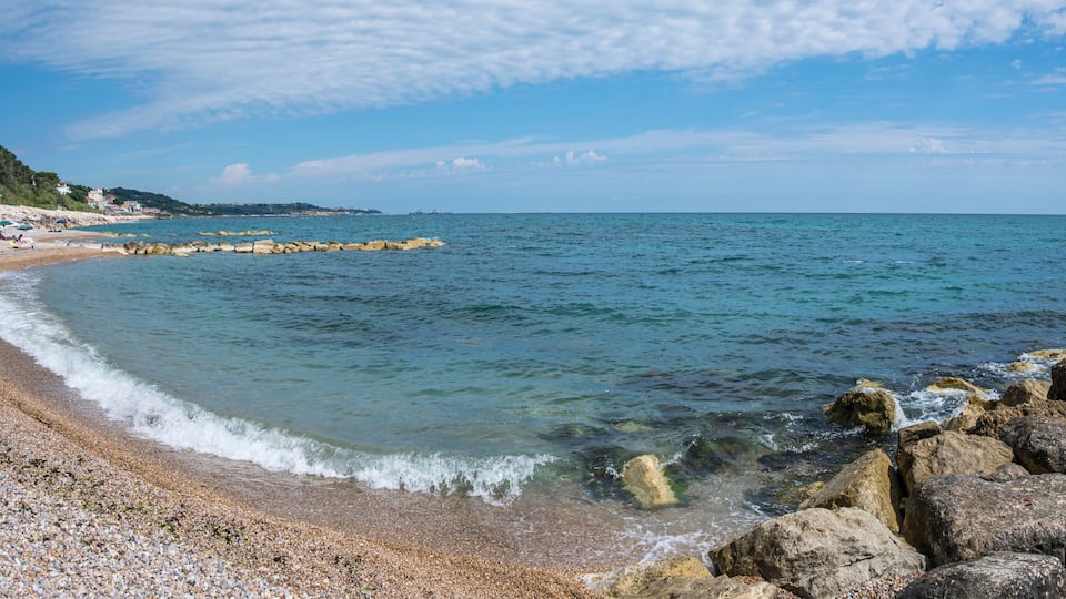 A pebble beach with clear water, a rocky breakwater, and a distant shoreline with buildings.