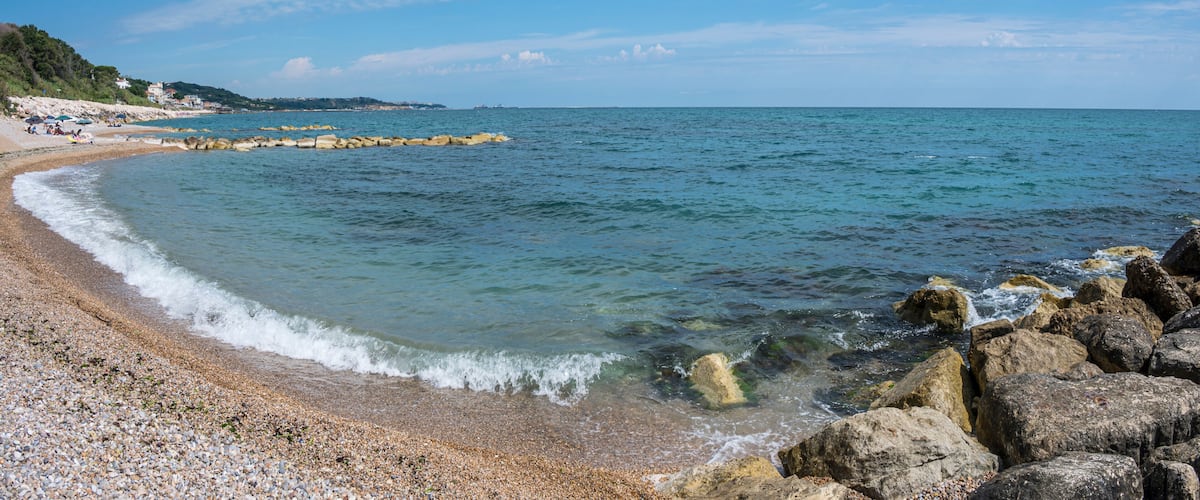 A pebble beach with clear water, a rocky breakwater, and a distant shoreline with buildings.