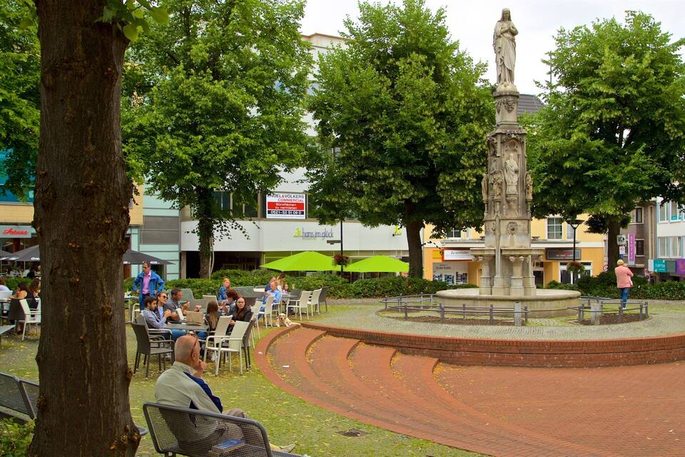 Een openbaar plein met een fontein in het midden, omringd door bomen en bankjes.