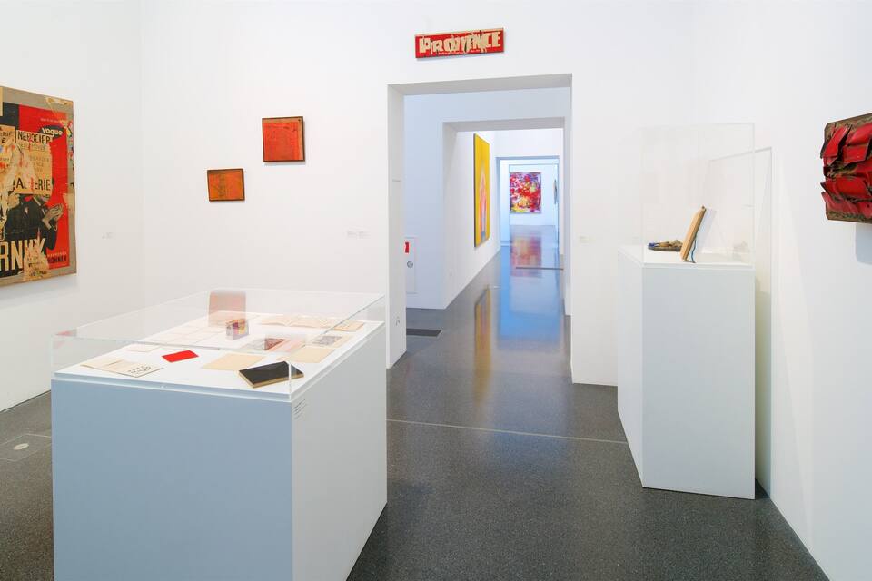 Una galeria d'art amb parets blanques i un sòl polit. Hi ha diverses obres d'art exposades, incloent un pòster gran i una escultura vermella.