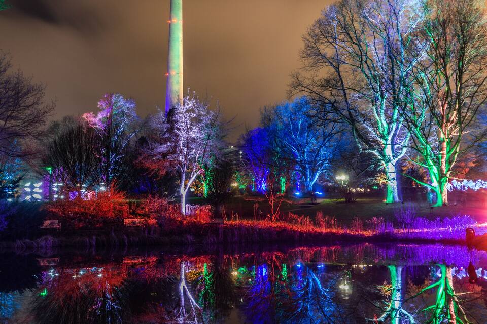 Uma torre com o topo iluminado, cercada por árvores decoradas com luzes coloridas, com os reflexos em um corpo d'água.