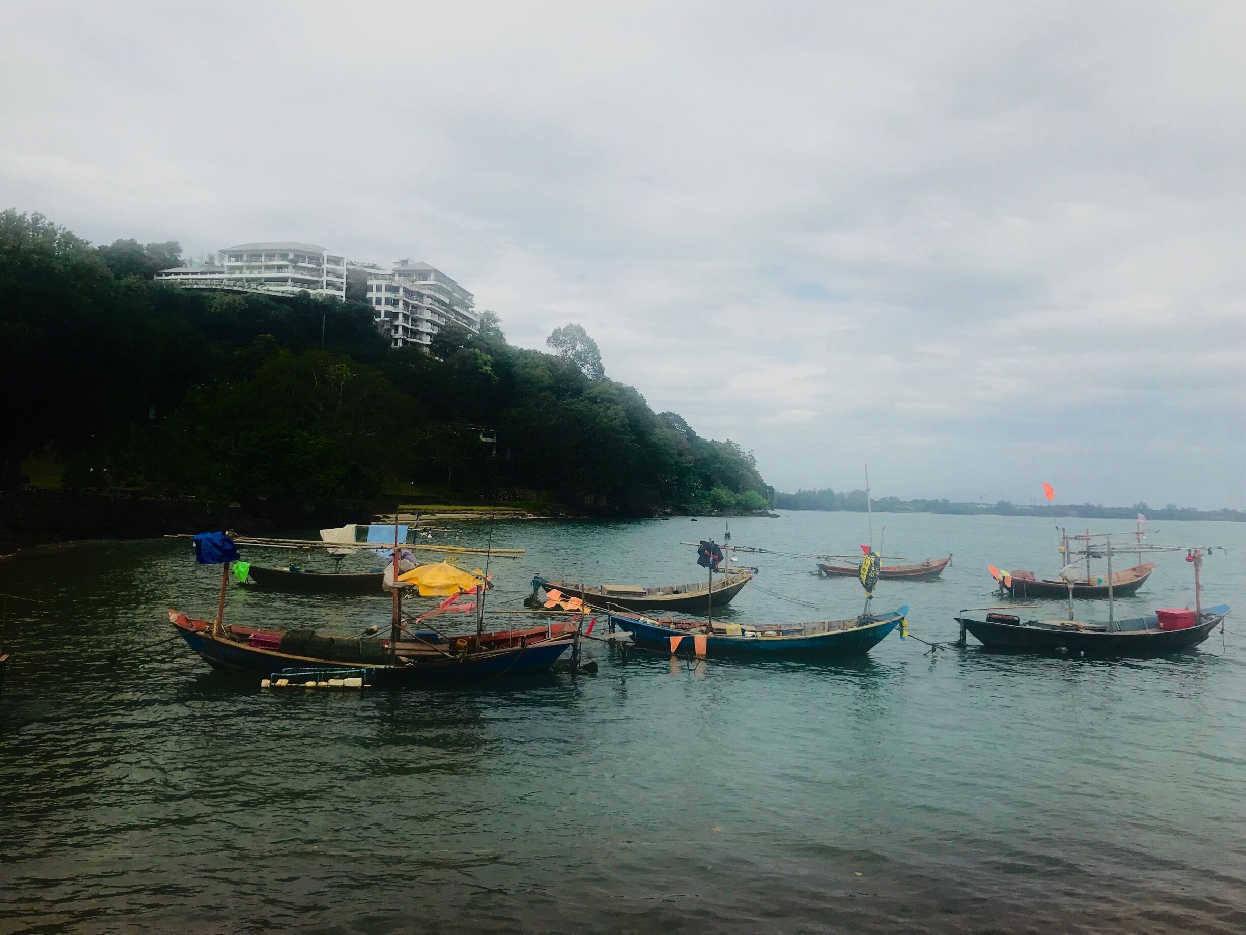 A calm river with several traditional boats docked along the shore. In the background, there is a modern building on a hill.