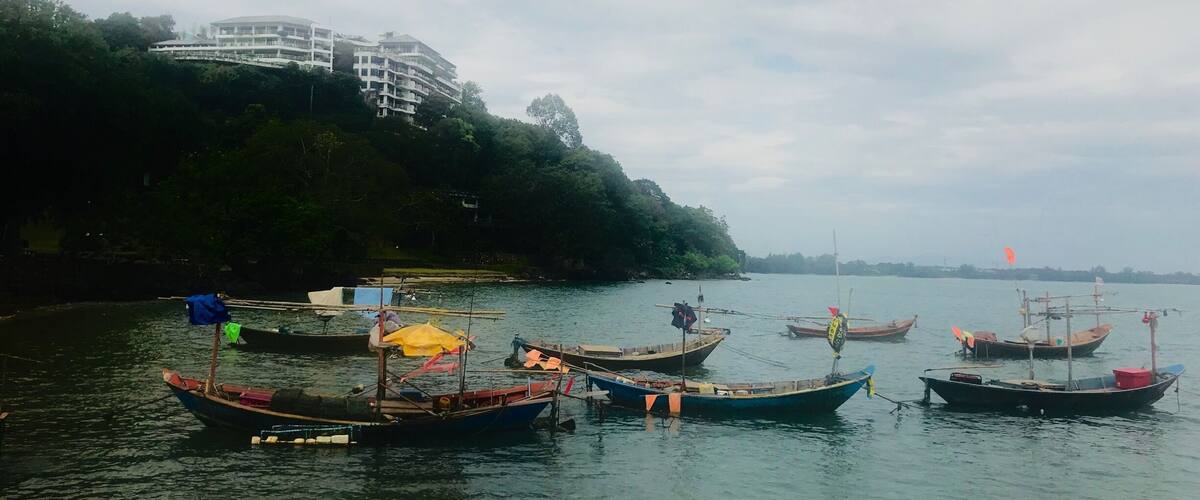 A calm river with several traditional boats docked along the shore. In the background, there is a modern building on a hill.