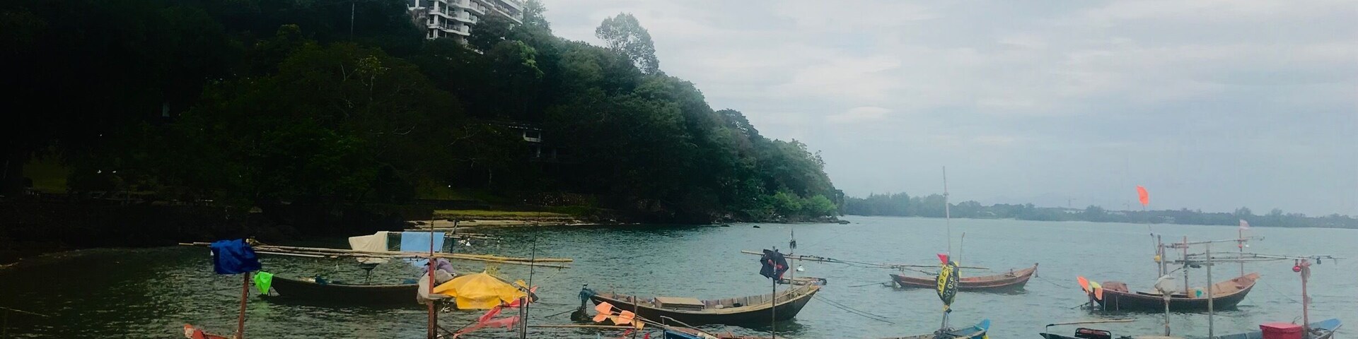 A calm river with several traditional boats docked along the shore. In the background, there is a modern building on a hill.