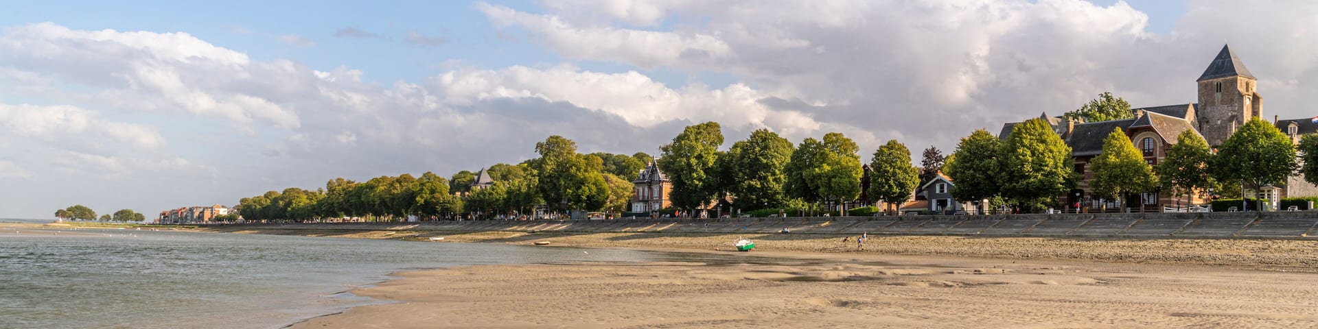 A sandy beach with gentle waves, a row of trees, and a historic building with a tower in the background.
