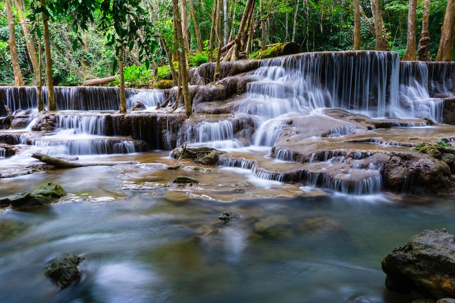 A multi-tiered waterfall in a forested area with rocky terrain and dense greenery.