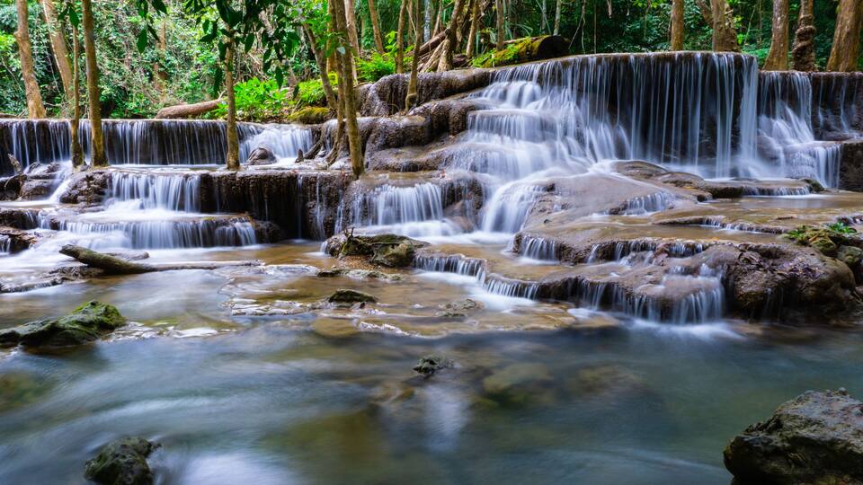 A multi-tiered waterfall in a forested area with rocky terrain and dense greenery.