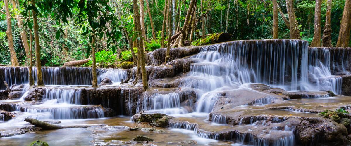 A multi-tiered waterfall in a forested area with rocky terrain and dense greenery.