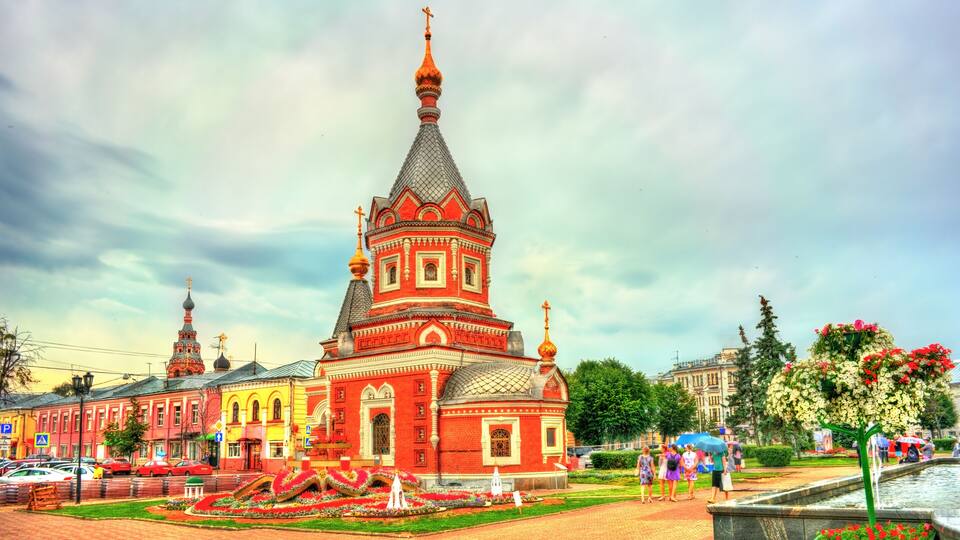 A red church with a tall spire and a bell tower, surrounded by a park with flowers and a fountain.