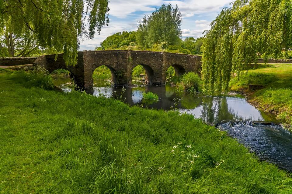 Uma ponte de pedra sobre um rio calmo, rodeada por vegetação exuberante e árvores.
