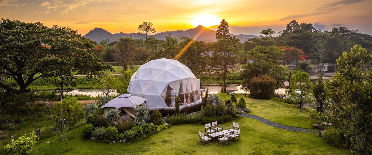 A white geodesic dome house surrounded by lush greenery and a clear sky.