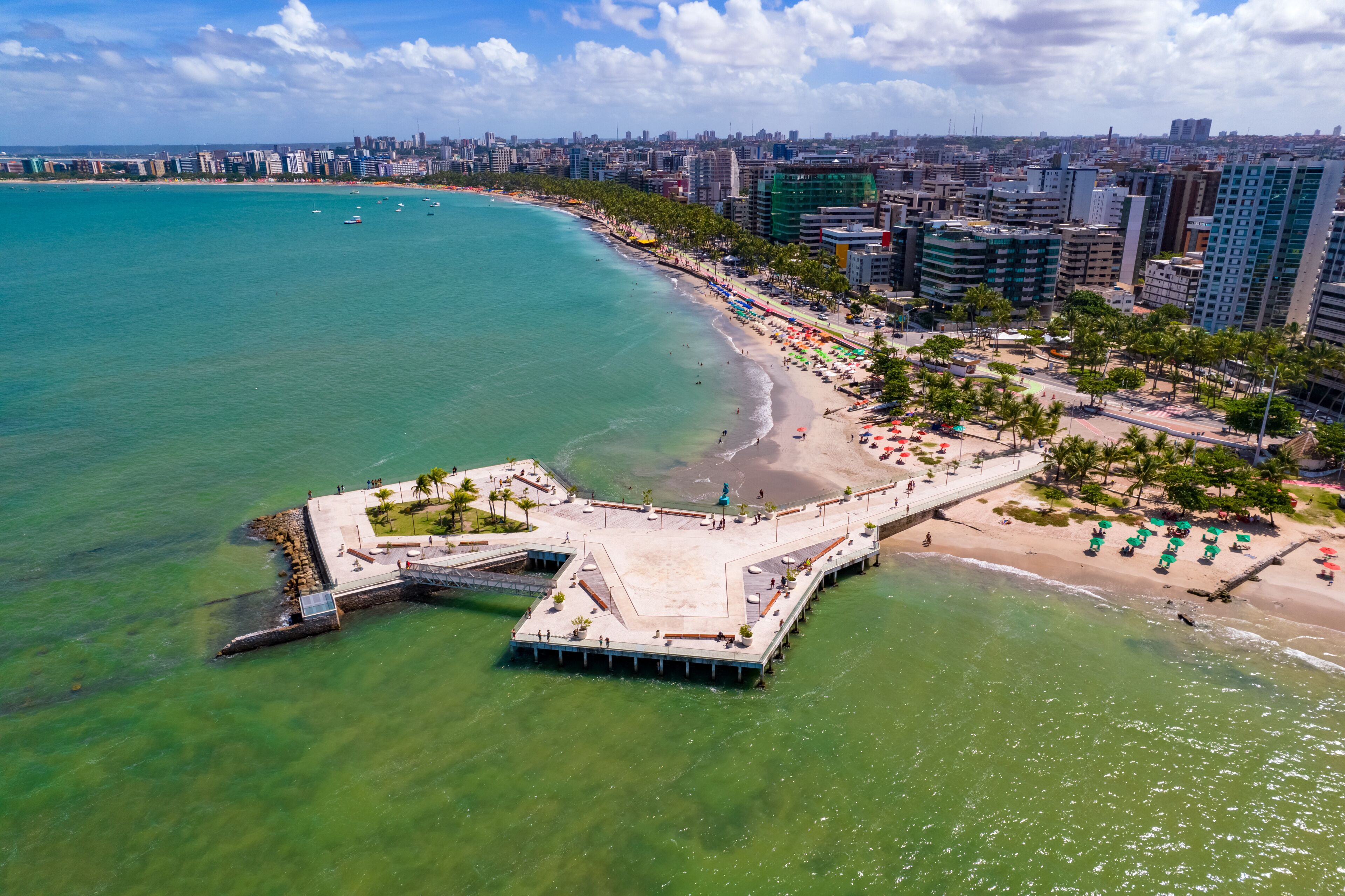 Un paysage urbain côtier avec une jetée imposante s’avançant dans la mer turquoise, une plage de sable bordée de palmiers et un décor urbain dense en arrière-plan.