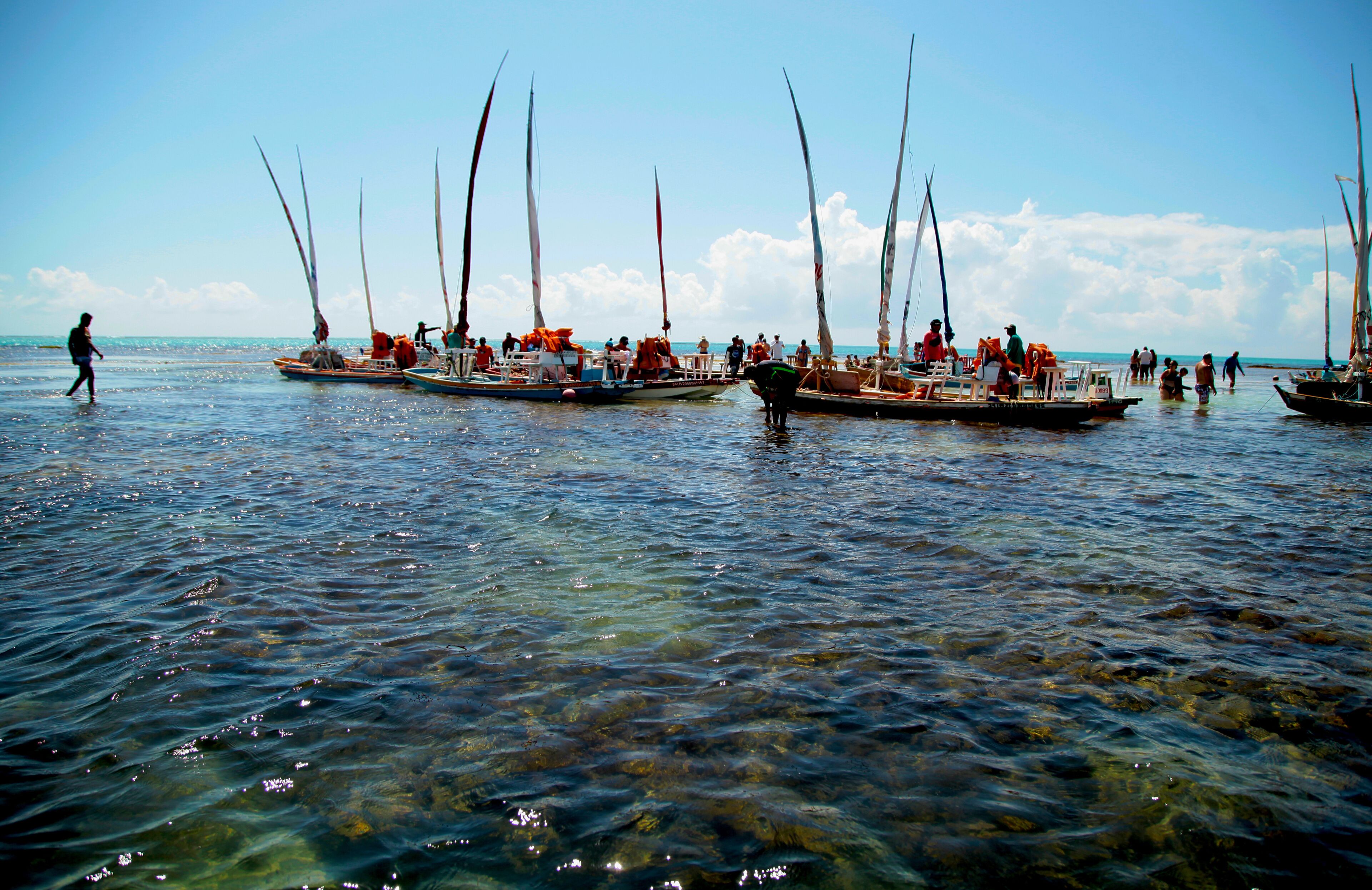 A calm sea with several sailboats anchored close to the shore, clear water, and a person walking along the water's edge.