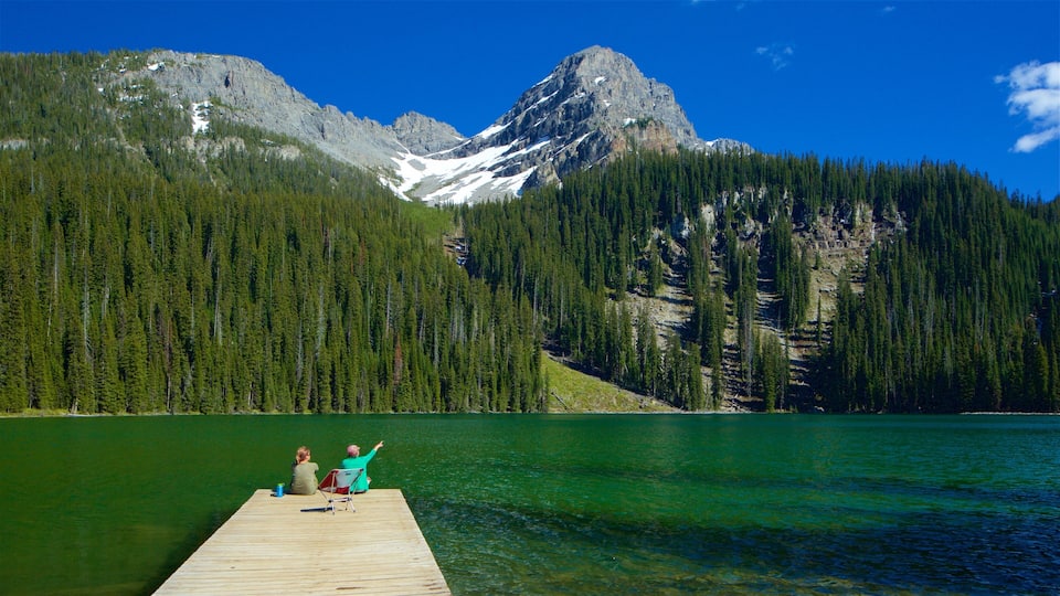 A wooden dock extending into a lake with two people sitting on it, surrounded by dense forests and a mountain range in the background.