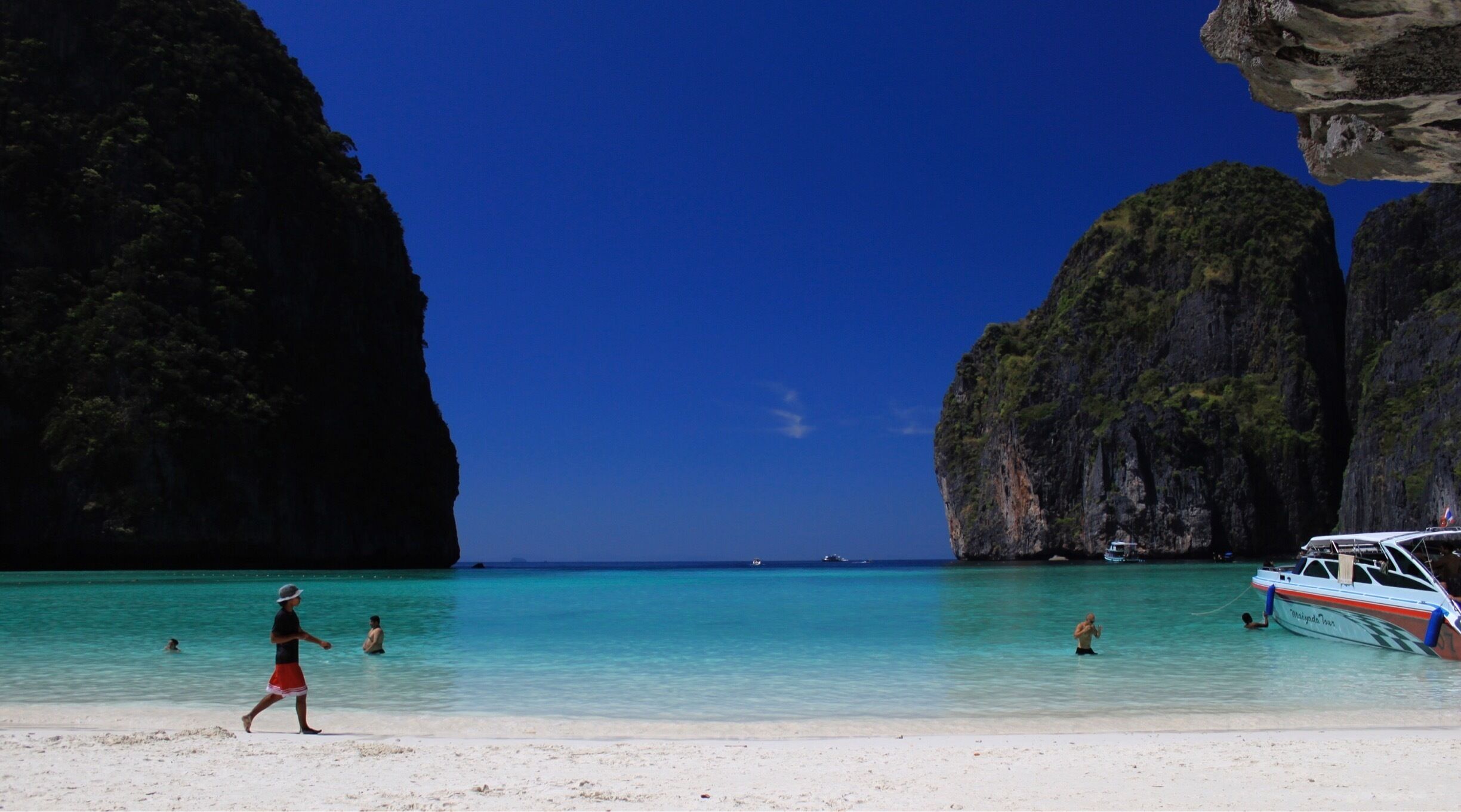 A beach with clear turquoise water, a boat named 'Myrinda Tour', and large limestone cliffs.