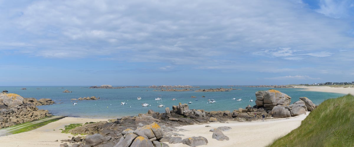A coastal landscape with a sandy beach, rocky outcrops, and calm sea with boats.