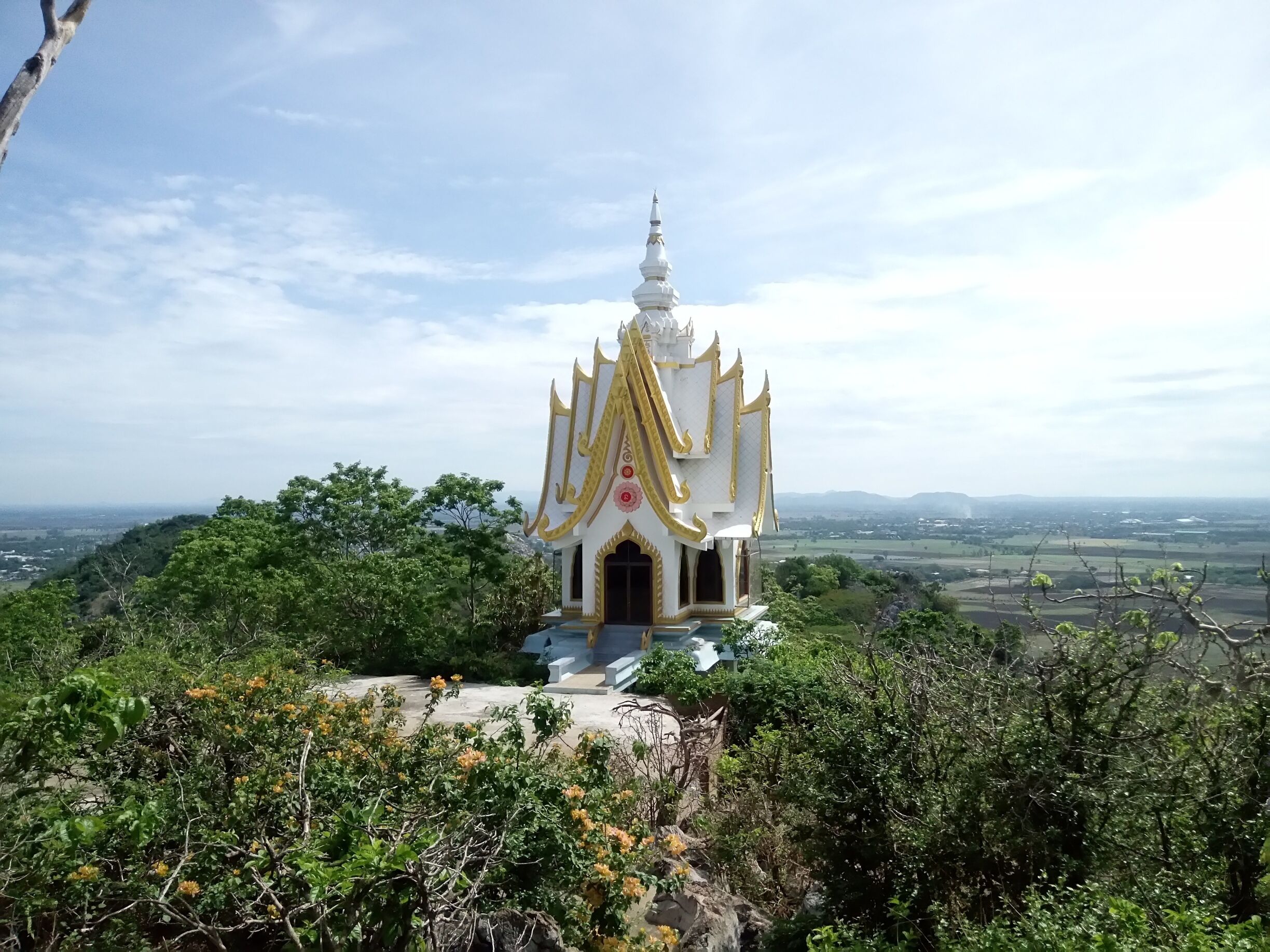 A white temple with golden accents surrounded by lush greenery and a distant landscape.
