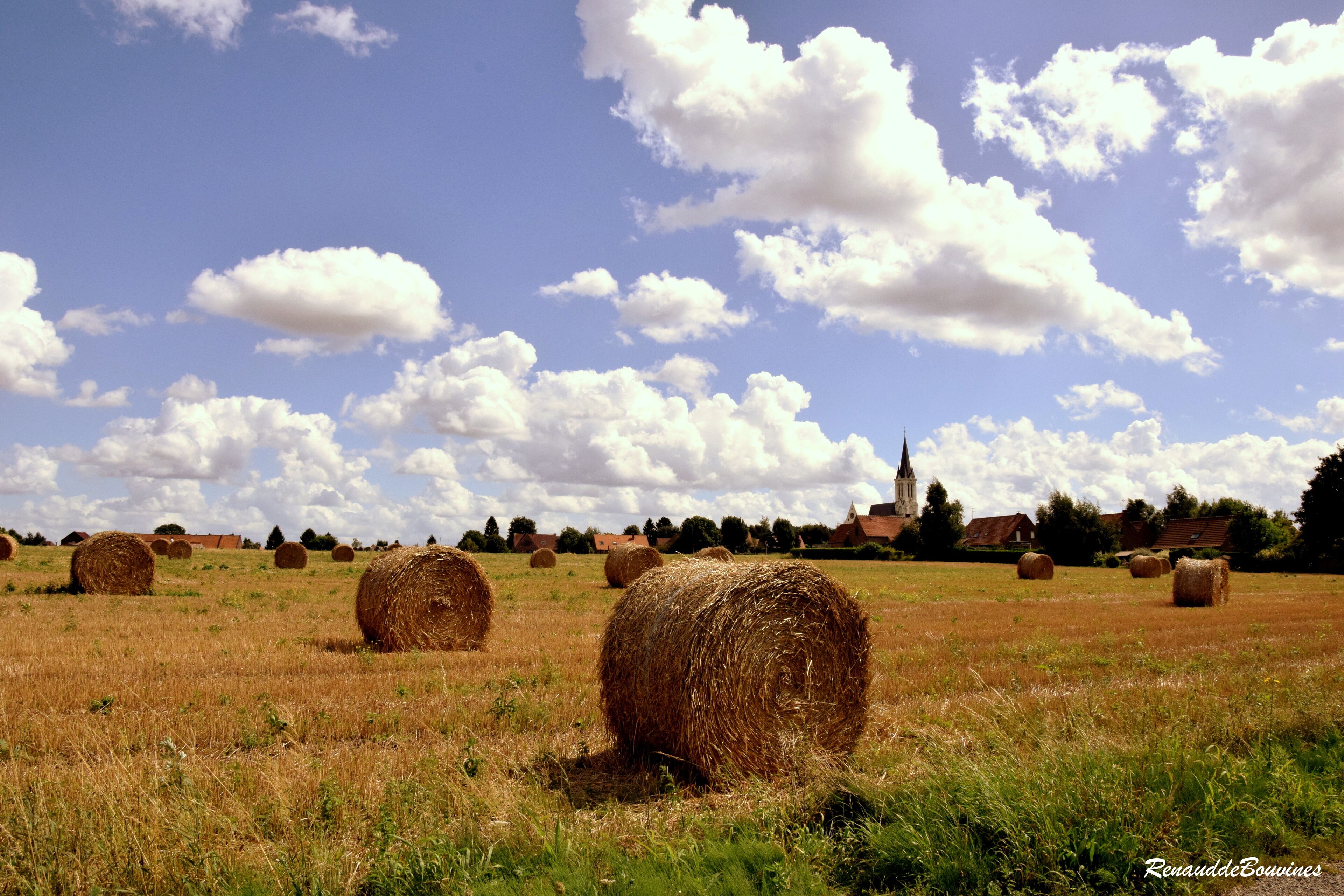 A field with hay bales, a church, and a clear sky with clouds.