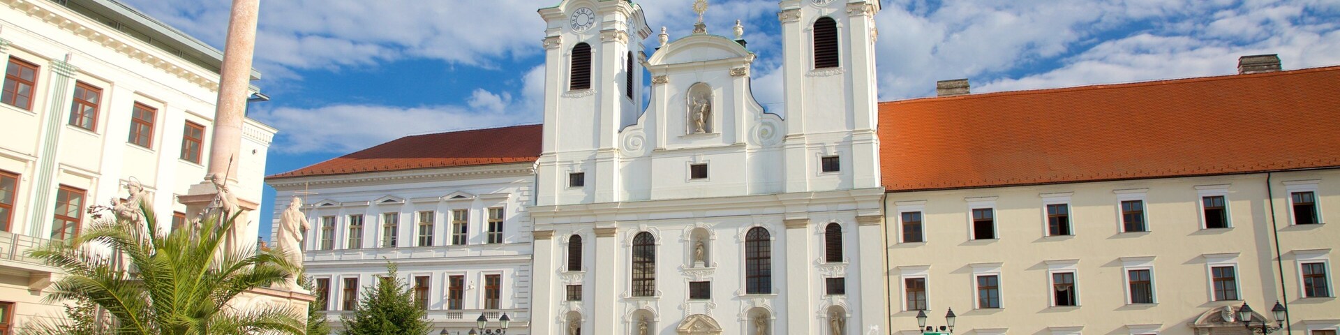 A historic plaza with a prominent church featuring two green domes, a tall column with a statue, and a fountain.