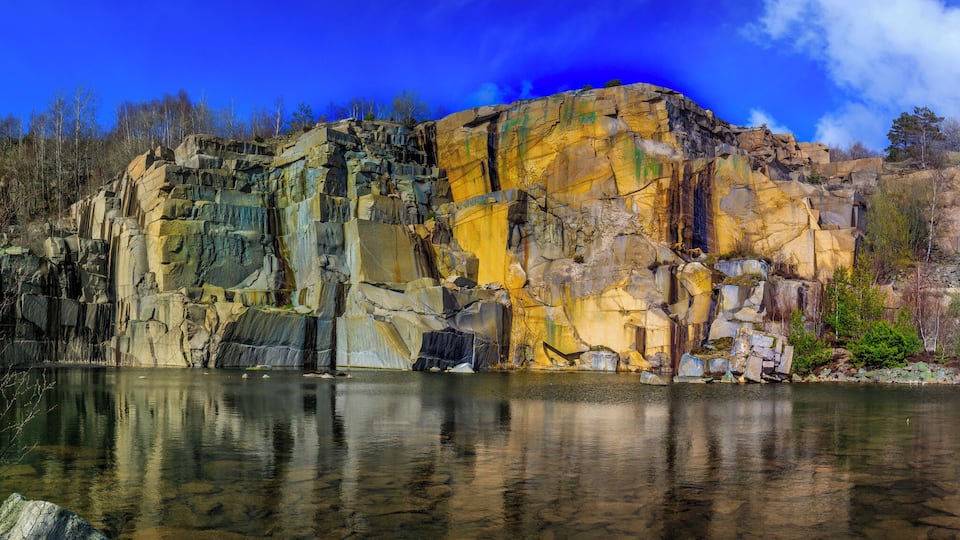 A rocky cliff with a clear reflection in a calm body of water, surrounded by trees and a blue sky.