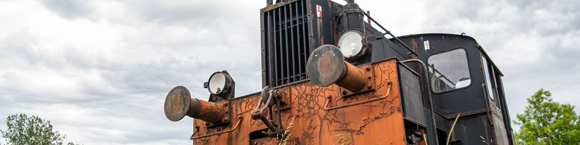 Eine verrostete Zuglokomotive in einem grasbewachsen Feld unter einem bewölkten Himmel.