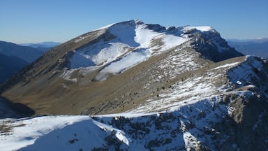 Pico nevado de montanha com encostas rochosas e cadeias montanhosas ao longe.
