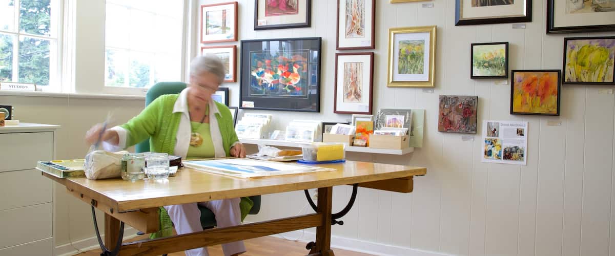 A room with a wooden table, various framed artworks on the wall, and a window with a view of trees.