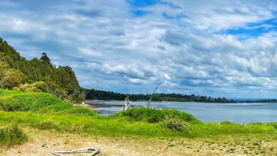 A coastal landscape with a grassy foreground, a bench, and a body of water under a partly cloudy sky.