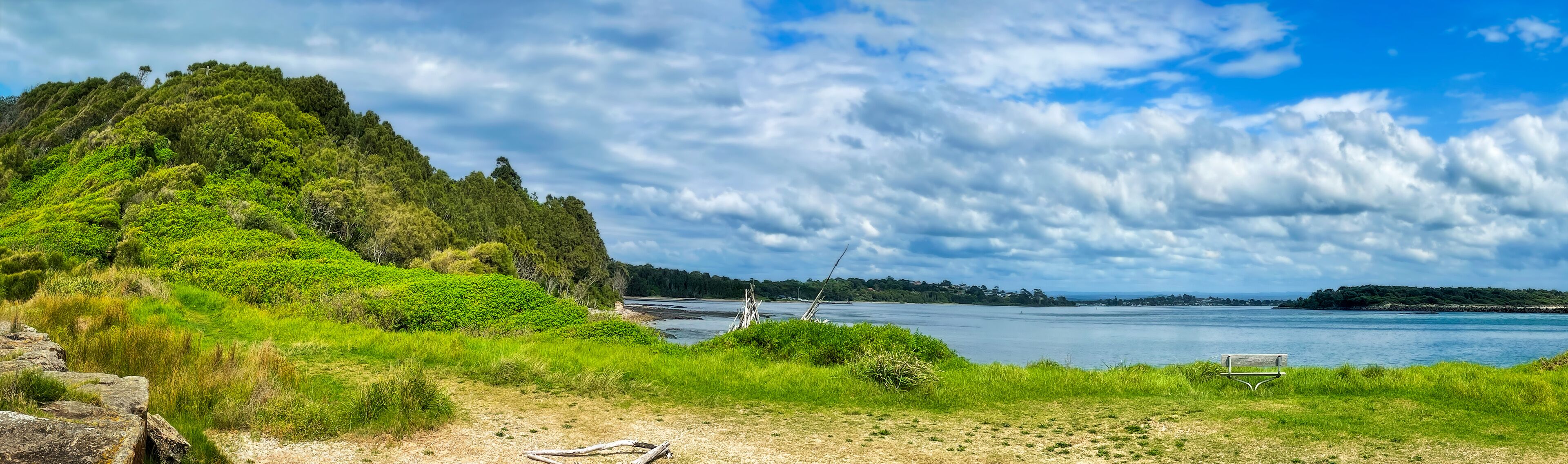 A coastal landscape with a grassy foreground, a bench, and a body of water under a partly cloudy sky.
