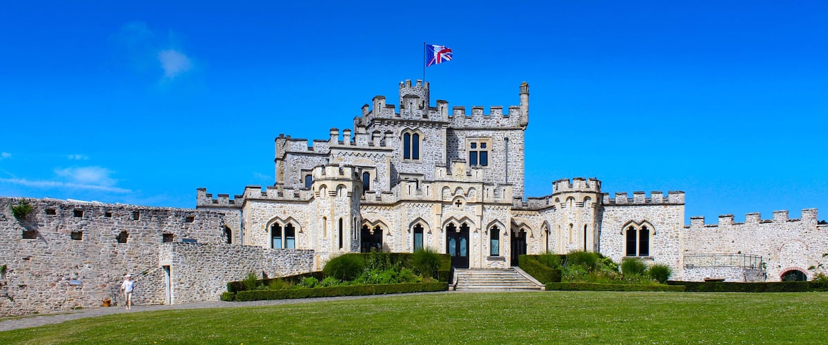 A large stone castle with a flag, surrounded by a well-maintained lawn and a stone wall.