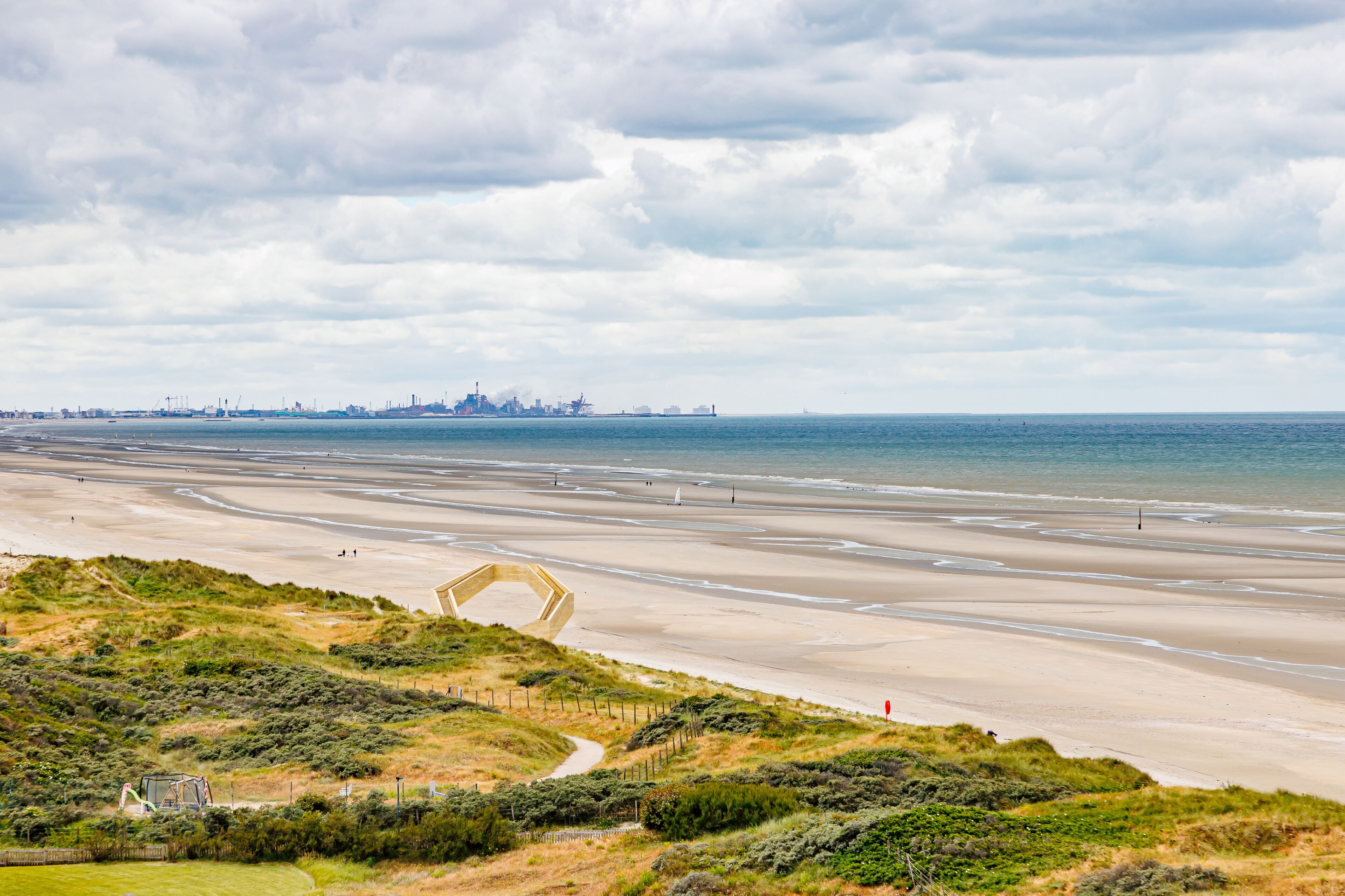 Panoramic landscape of beach with North Sea and industrial complex in Dunkirk, France in background, stormy sky covered with clouds, viewpoint and dunes, cloudy day De Panne, West Flanders, Belgium