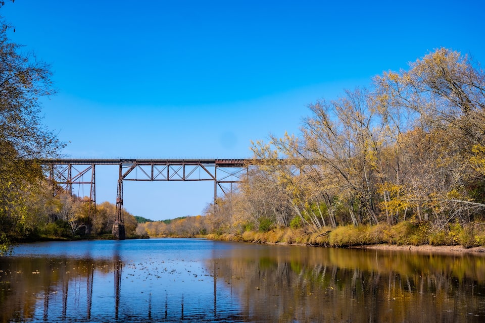 Eine Stahlbrücke über einem ruhigen Fluss, umgeben von Herbstbäumen.