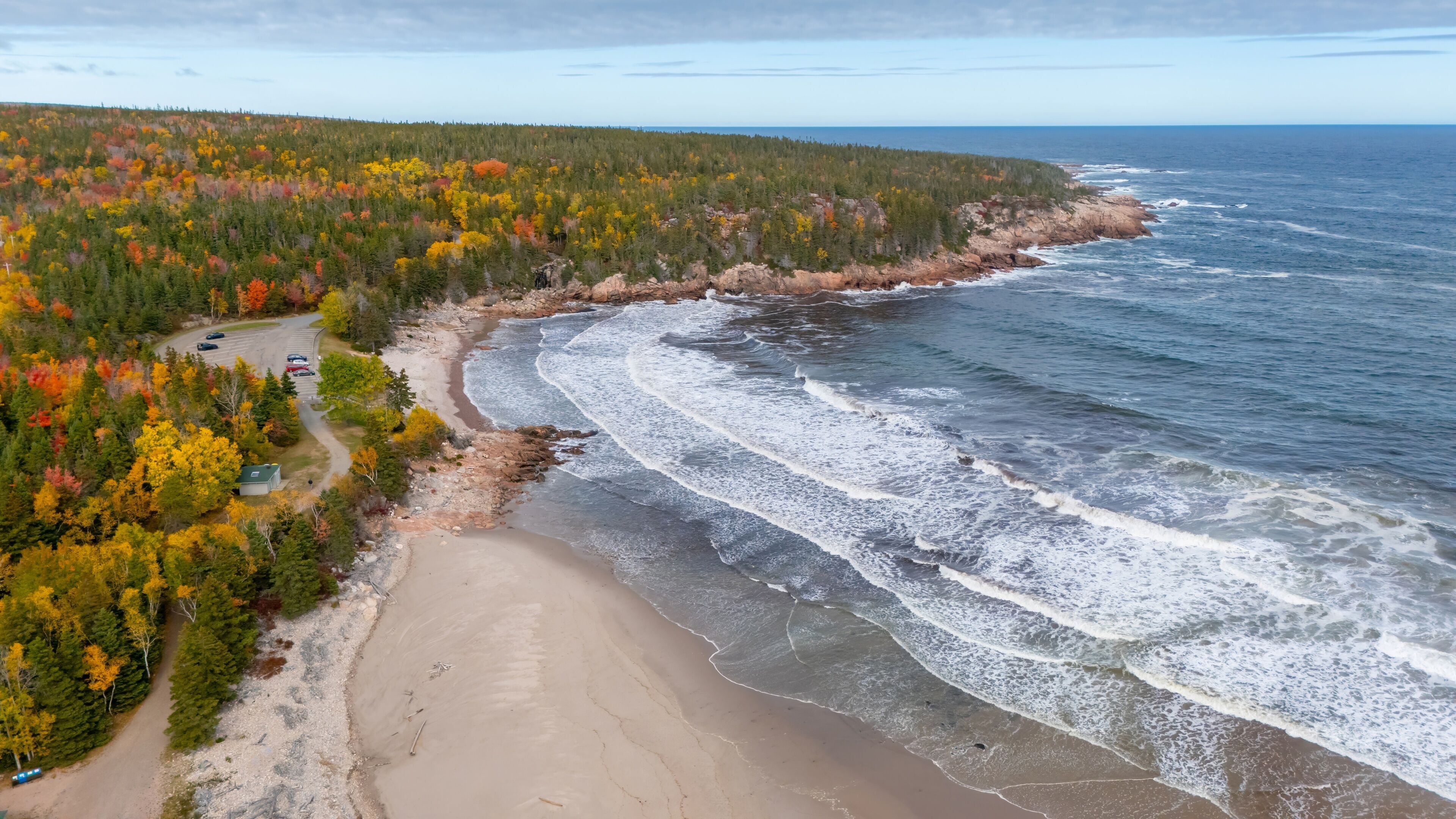 A coastal landscape with a sandy beach, a rocky shoreline, and a forested area with autumn-colored trees.