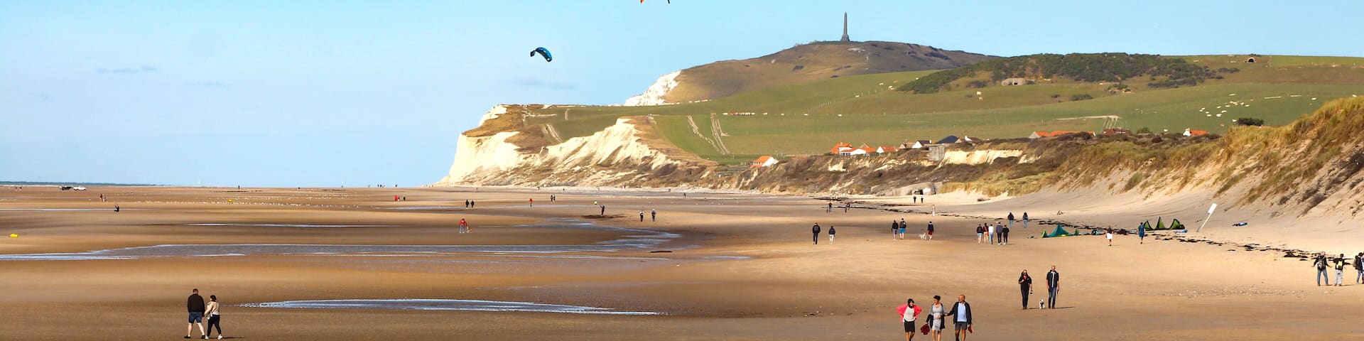 A wide sandy beach with people walking and kites flying.